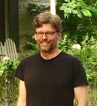 Mike Bicknell, wearing a black T-shirt, stands in front of a background of trees and vines in front of a cream-colored stone house.
