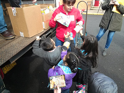 Three children standing behind a truck are handing food boxes and can to an adult.